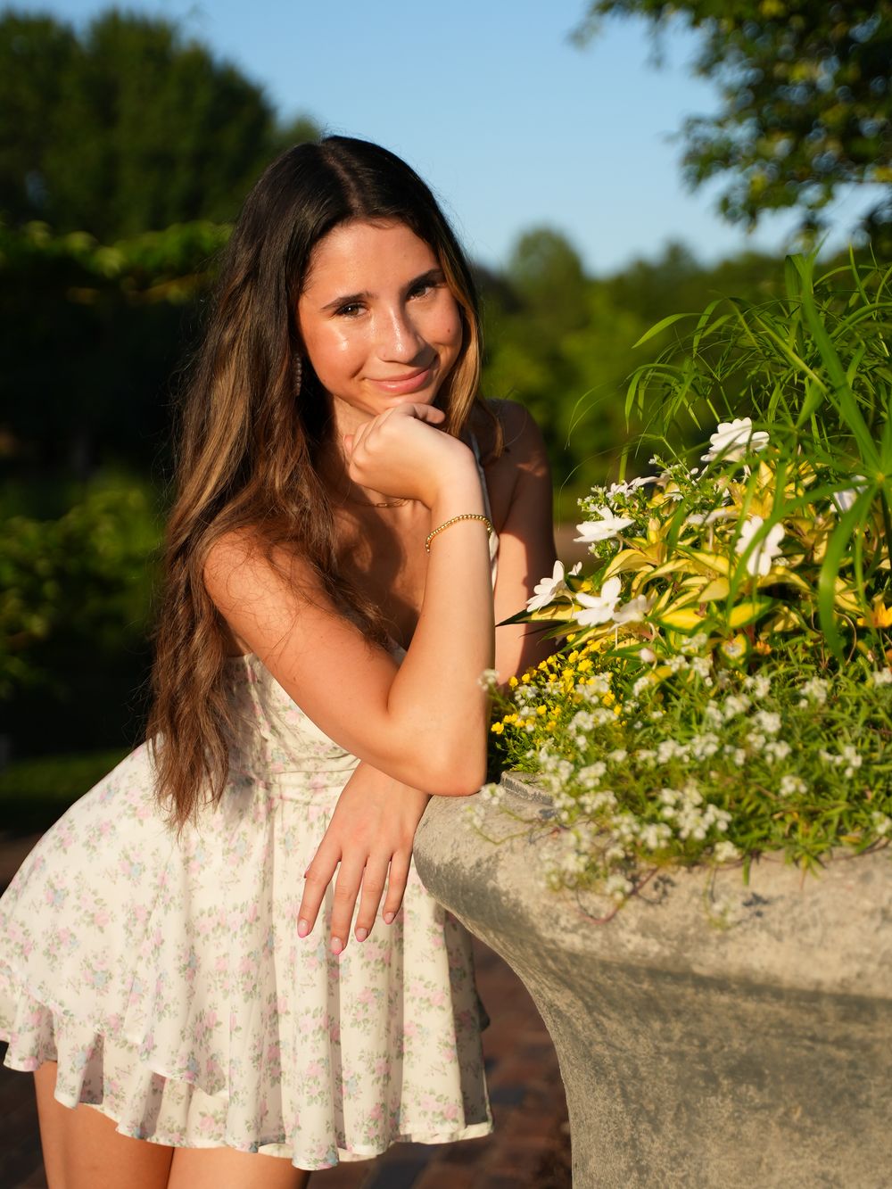 Young woman in floral sundress smiling beside a stone planter with white and yellow flowers in golden hour light.