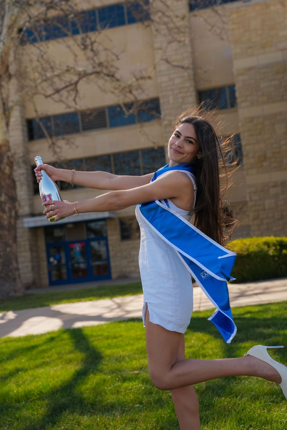 Young woman in white dress with blue graduation sash poses outdoors holding champagne bottle on one leg.
