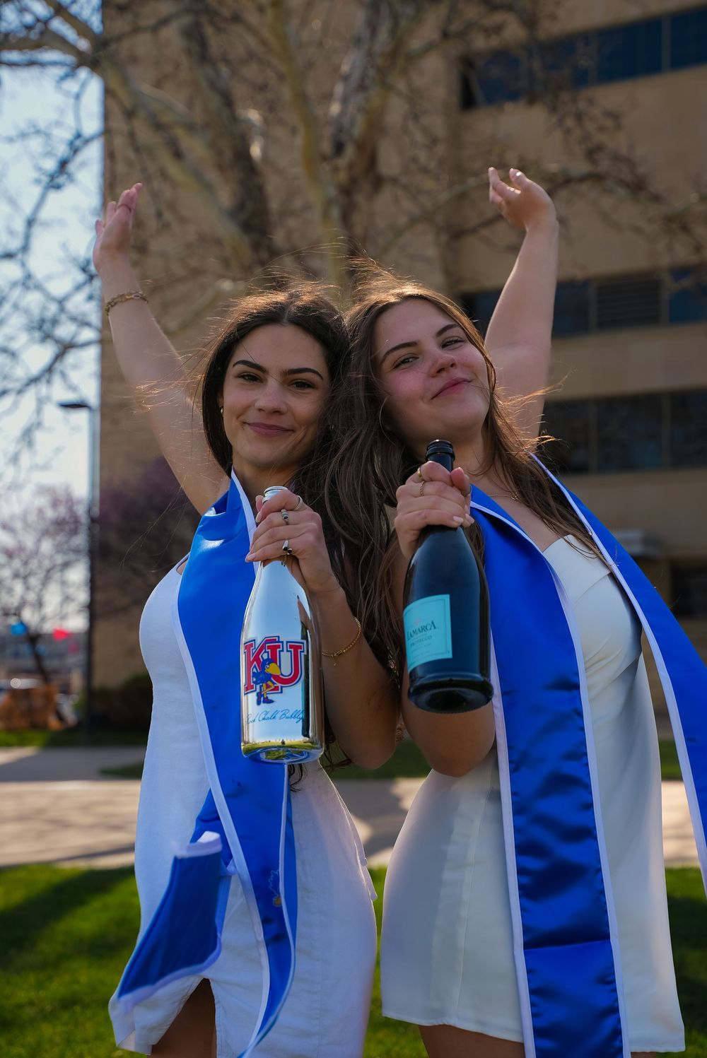Two smiling women in white dresses with blue graduation sashes celebrate outdoors holding champagne bottles.