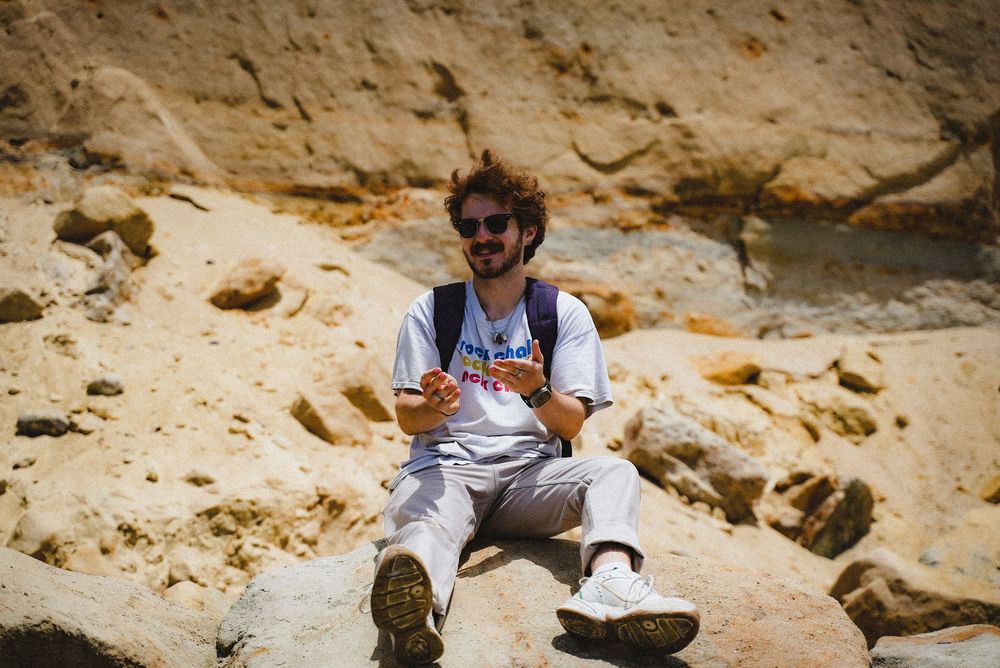 Man with curly hair and sunglasses sitting on rocky terrain, wearing a white t-shirt and light pants, smiling outdoors.