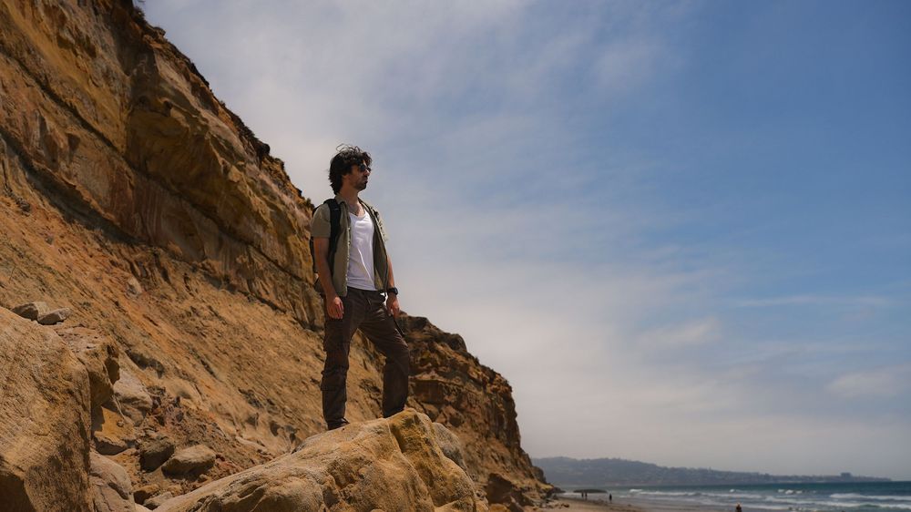 Man standing on rocky coastal cliff overlooking the ocean under a partly cloudy blue sky.