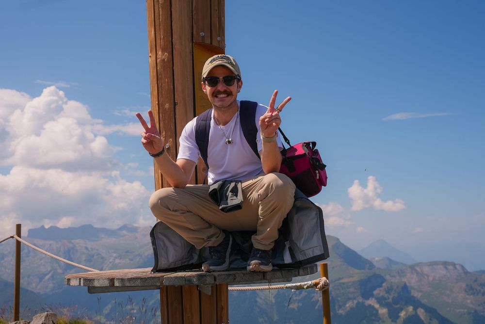 Hiker sitting on mountain summit cross making peace signs with stunning alpine panorama and blue sky.