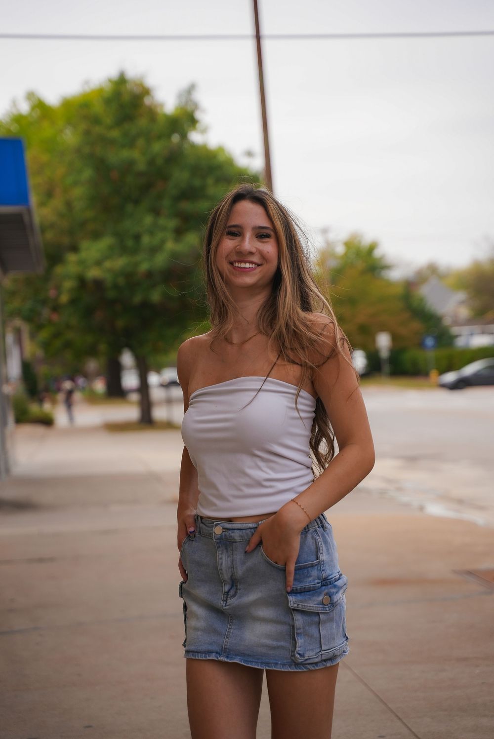 Young woman smiling in white tube top and denim cargo mini skirt, posing on a sidewalk with trees in the background.
