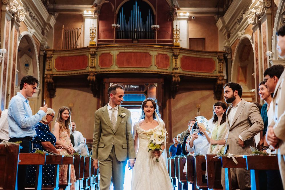 Bride and groom walk down the aisle of an ornate church, surrounded by smiling guests during a wedding ceremony.