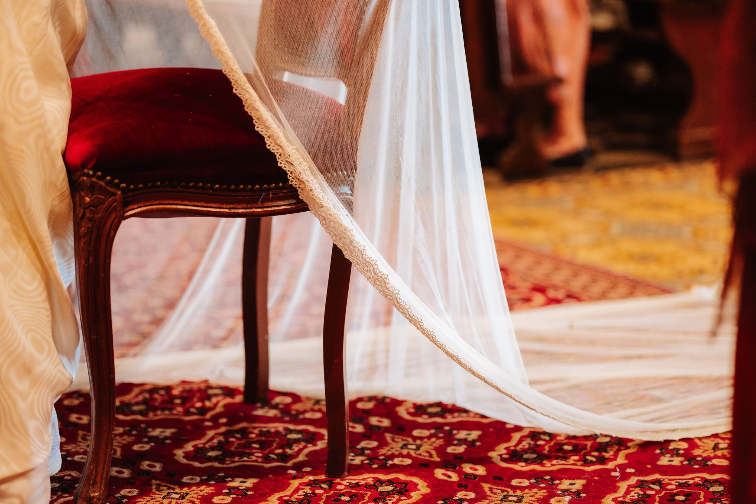 Elegant wedding veil draped over an ornate wooden chair on a red patterned carpet in a ceremonial venue.