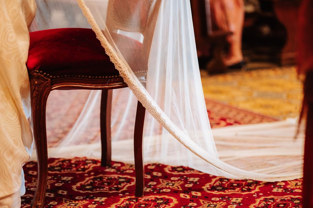 Elegant wedding veil draped over an ornate wooden chair on a red patterned carpet in a ceremonial venue.