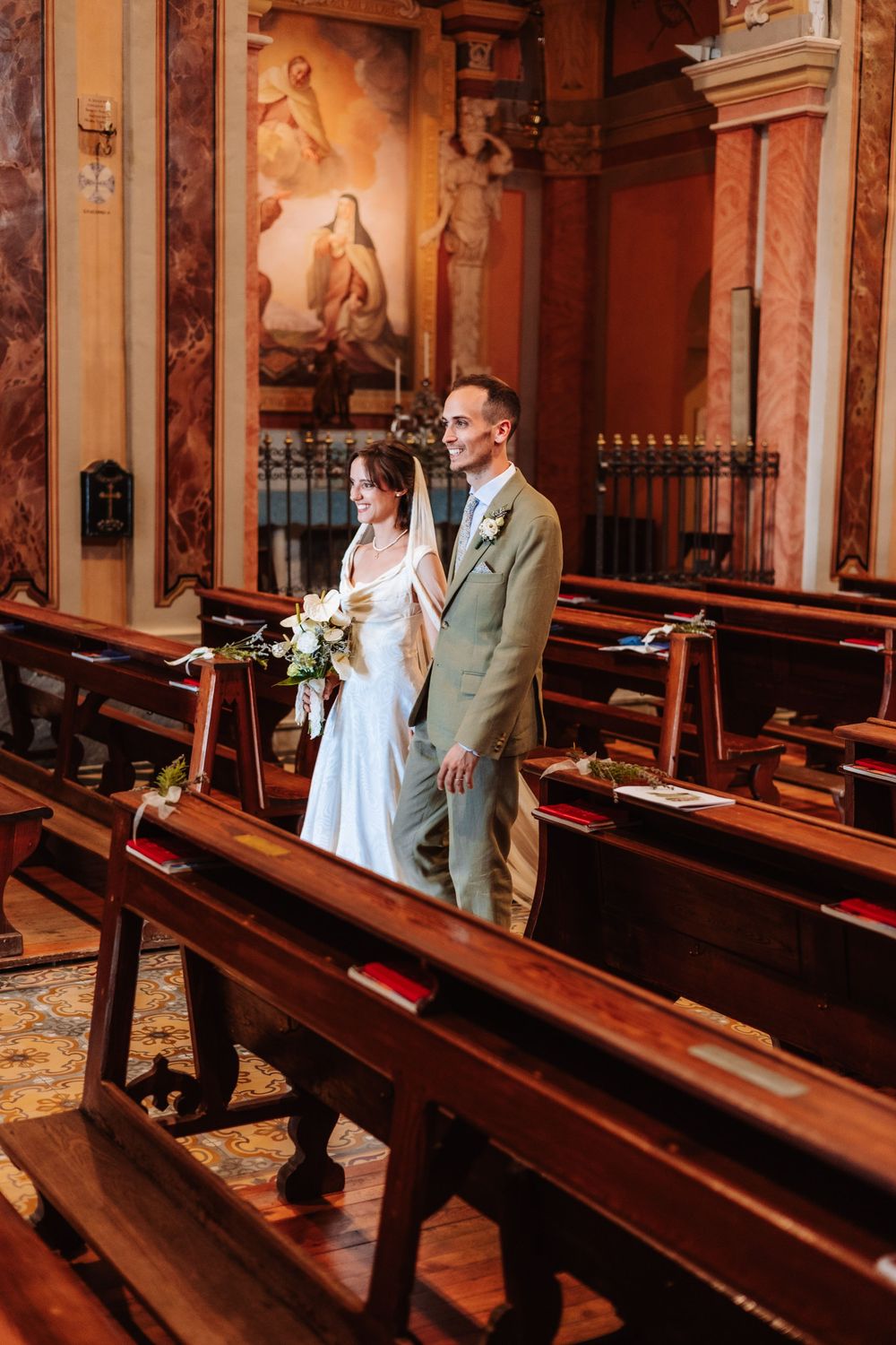 Bride and groom walking down the aisle of an ornate church with wooden pews and baroque paintings.
