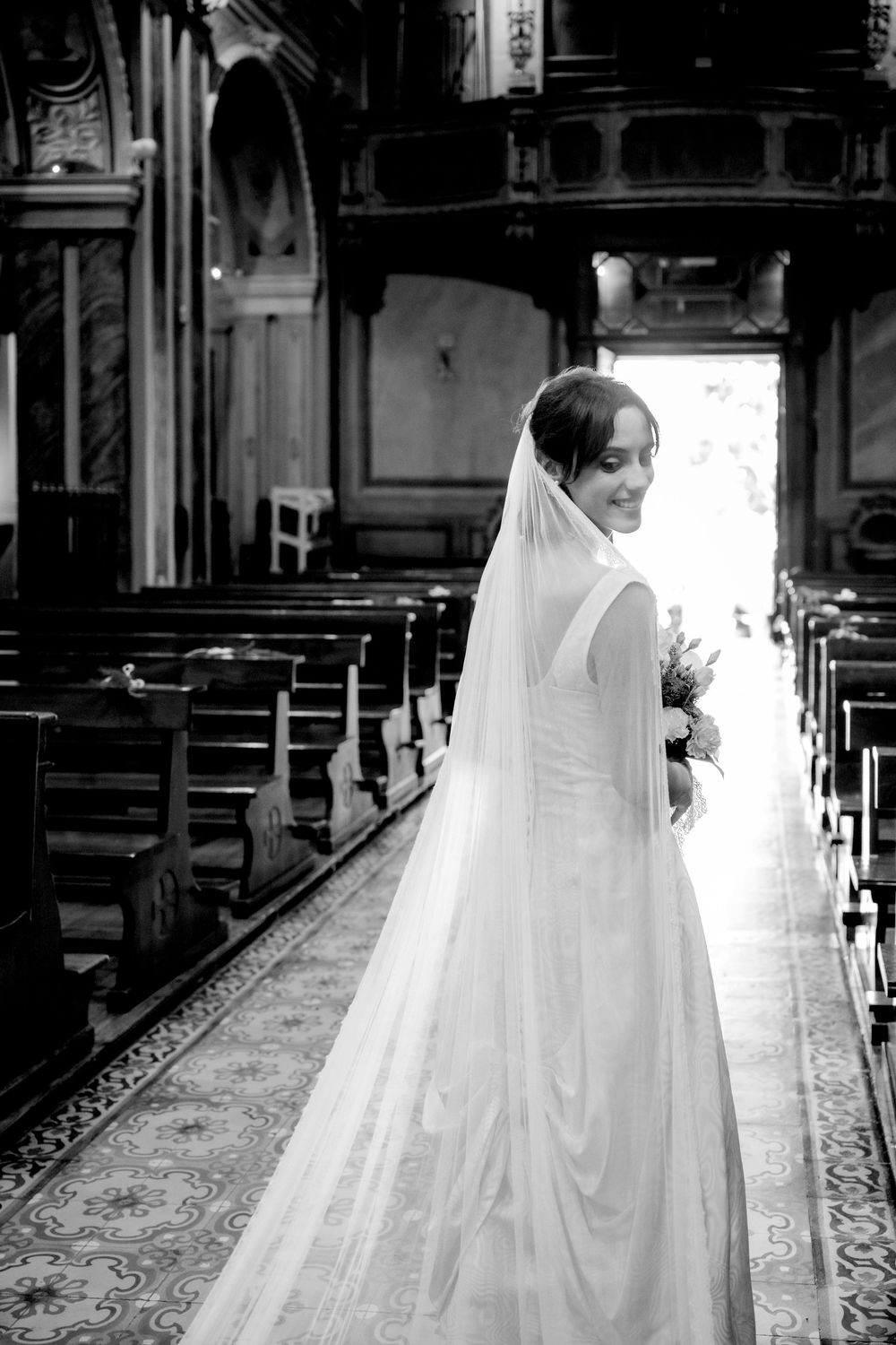 Bride in white gown and veil smiling in church aisle, holding bouquet, black and white wedding photo.