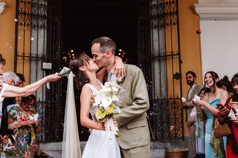 Newlyweds share a kiss outside a church door while guests celebrate with confetti and applause at their wedding.