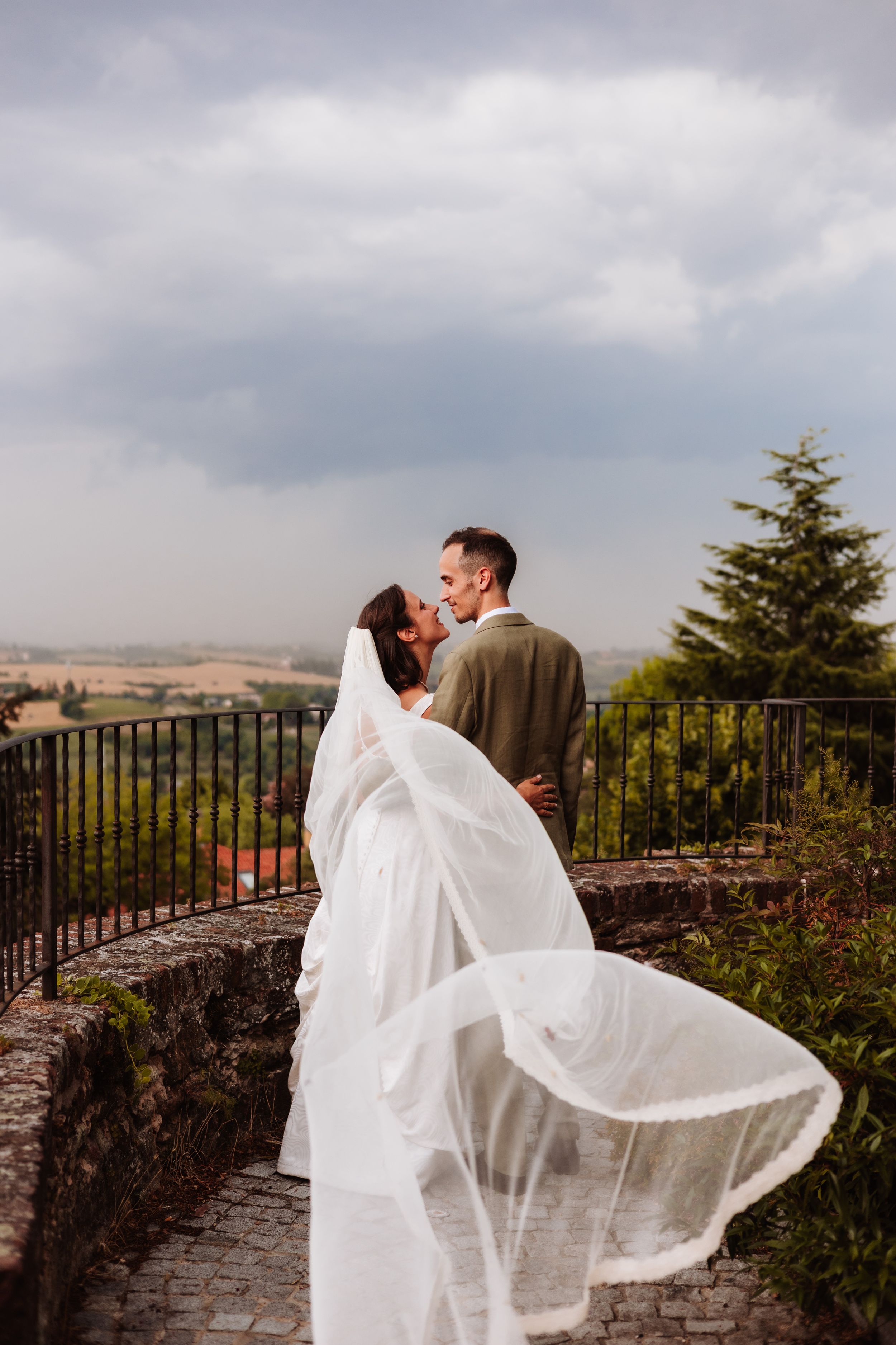 Bride and groom sharing a romantic kiss on a scenic overlook, with her veil flowing in the wind under a cloudy sky.