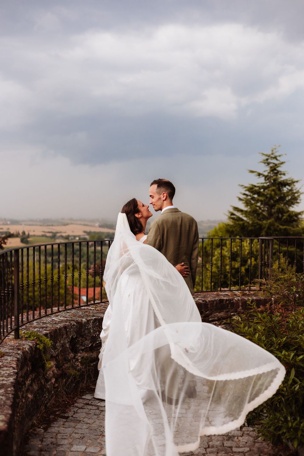 Bride and groom sharing a romantic kiss on a scenic overlook, with her veil flowing in the wind under a cloudy sky.