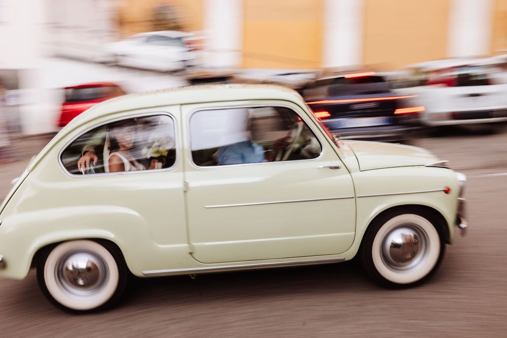 Vintage cream Fiat 600 car driving through city streets, captured with motion blur effect.
