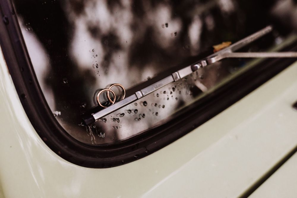 Wedding rings resting on a vintage car window wiper, with rain droplets on the glass in a romantic outdoor setting.