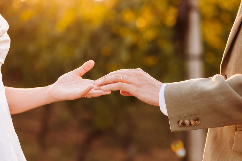 Two people gently touching hands outdoors during a wedding ceremony with warm golden autumn bokeh background.