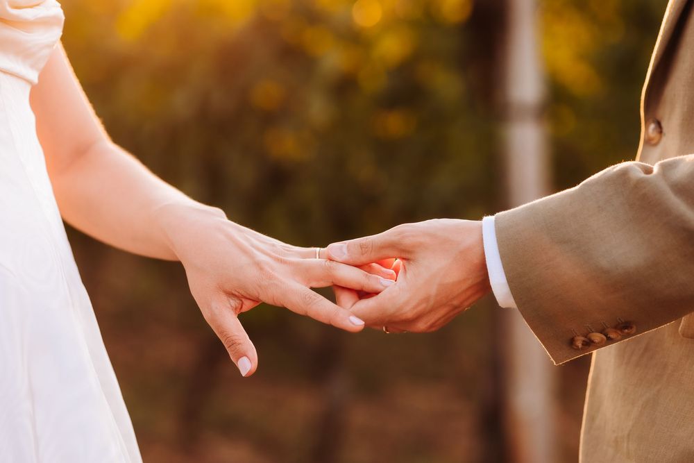 Close-up of a couple exchanging wedding rings outdoors with warm golden bokeh background.