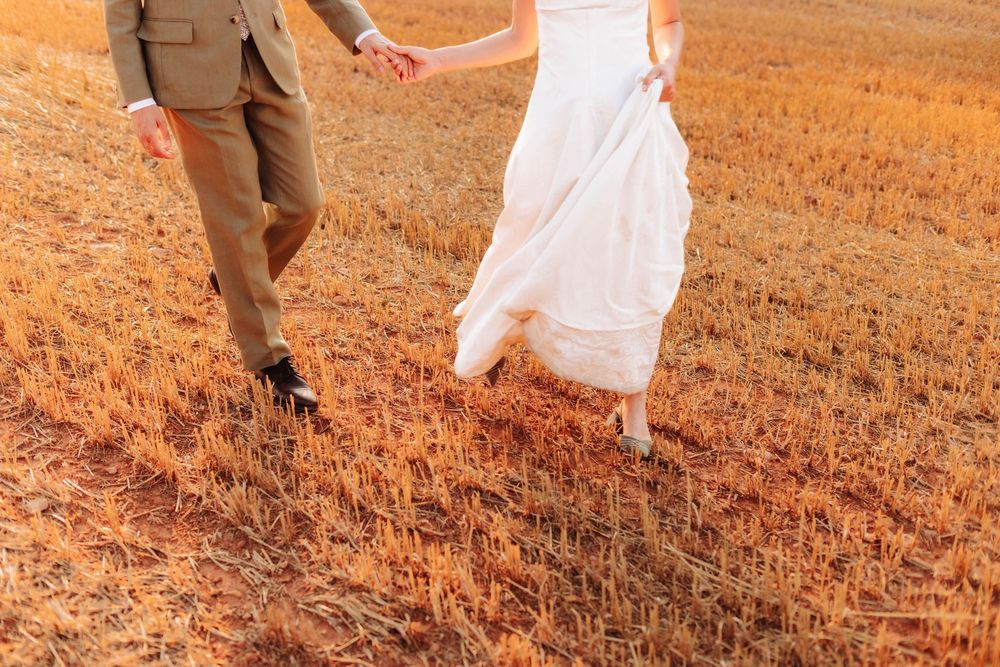 Bride and groom holding hands walking through golden wheat field at sunset in wedding attire.