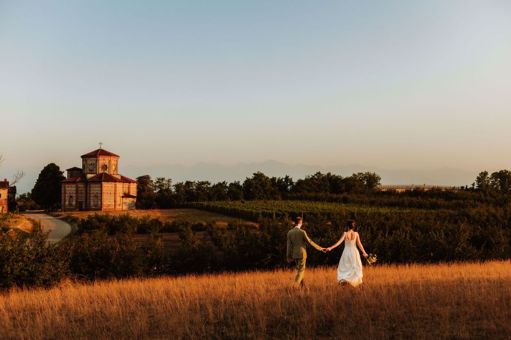 Couple holding hands walking through golden fields at sunset with a historic Tuscan villa and vineyards in the background.