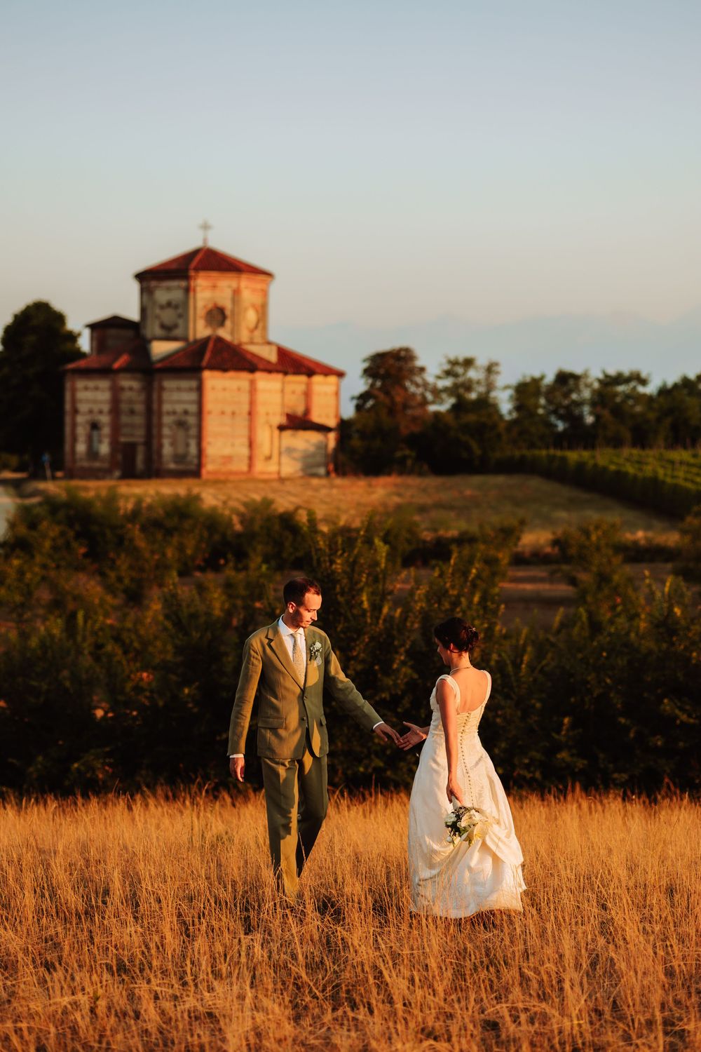 Bride and groom hold hands in golden field at sunset, with a historic red brick chapel and vineyard in the background.