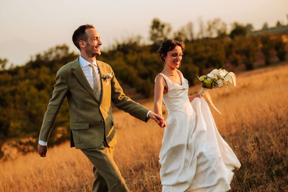 A joyful bride and groom walk hand-in-hand through a golden field at sunset on their wedding day.