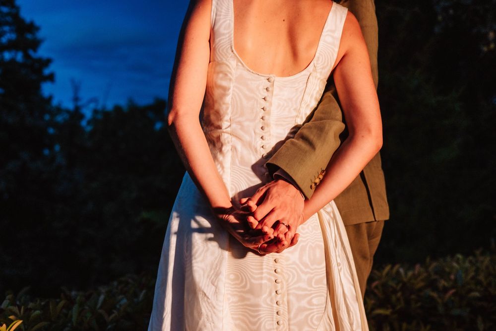Couple holding hands at dusk, bride in white button-back dress with groom in suit jacket outdoors.