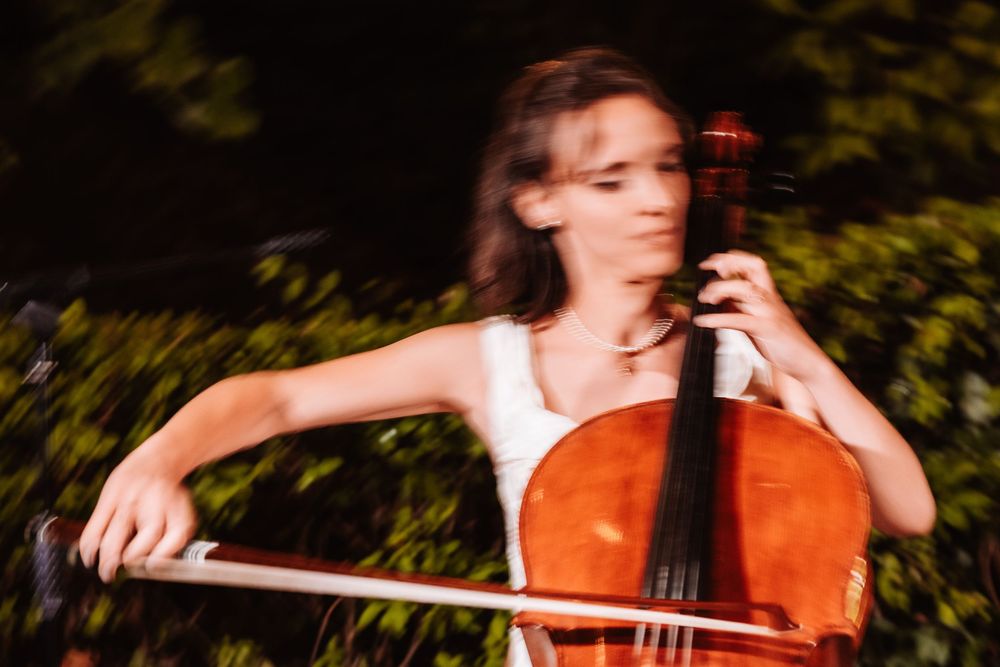 Young woman playing cello outdoors, motion blur effect, green foliage background, wearing white top.