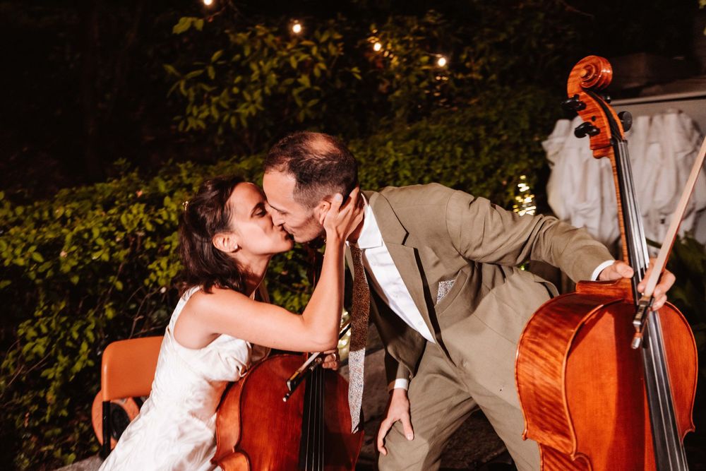 Bride and groom sharing a romantic kiss between two cellos at an outdoor evening wedding reception.