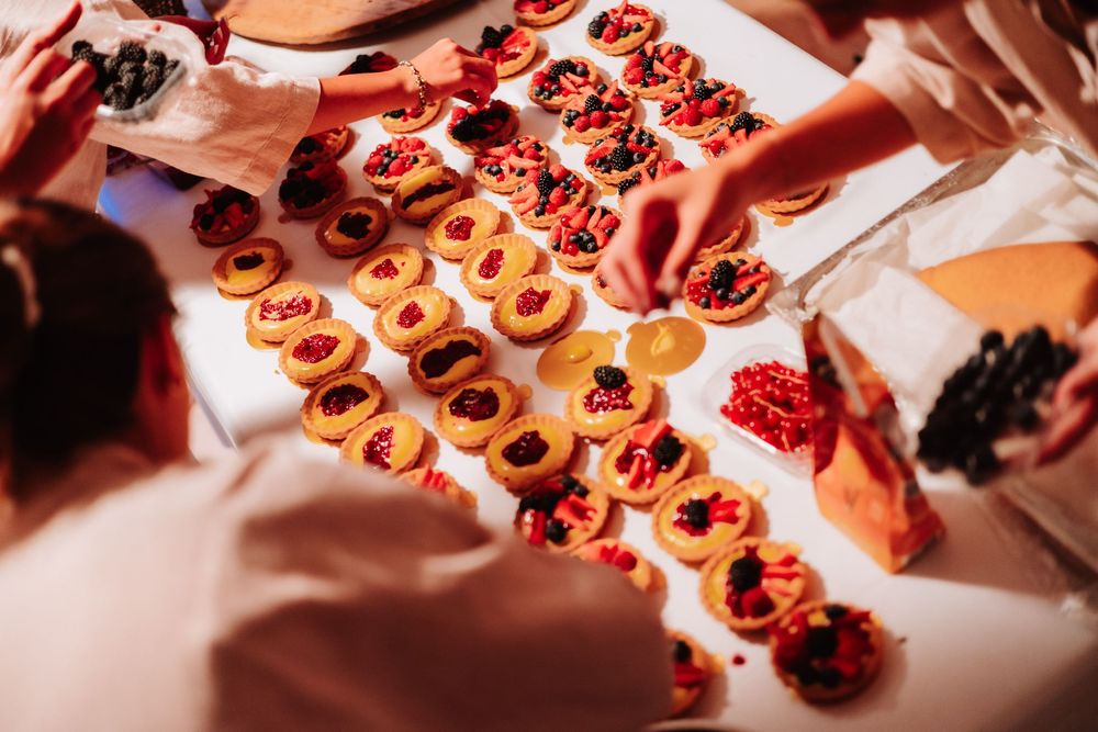 Overhead view of colorful fruit tarts with berries arranged on a white table being prepared by catering staff.