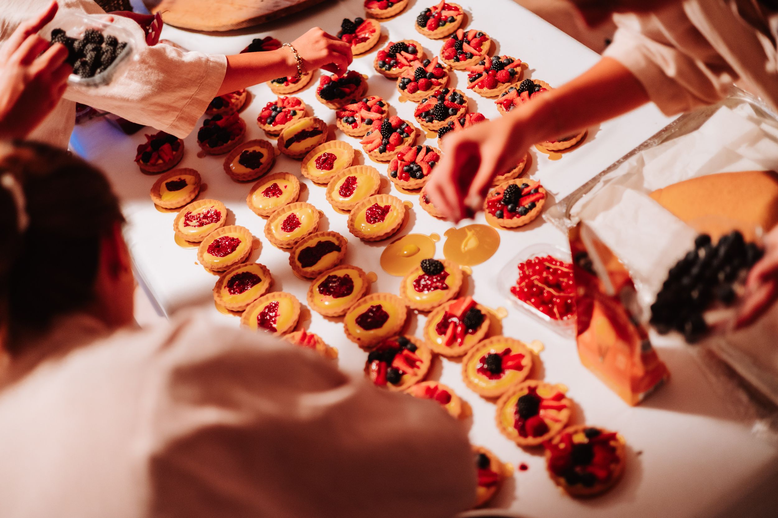 Overhead view of colorful fruit tarts with berries arranged on a white table being prepared by catering staff.
