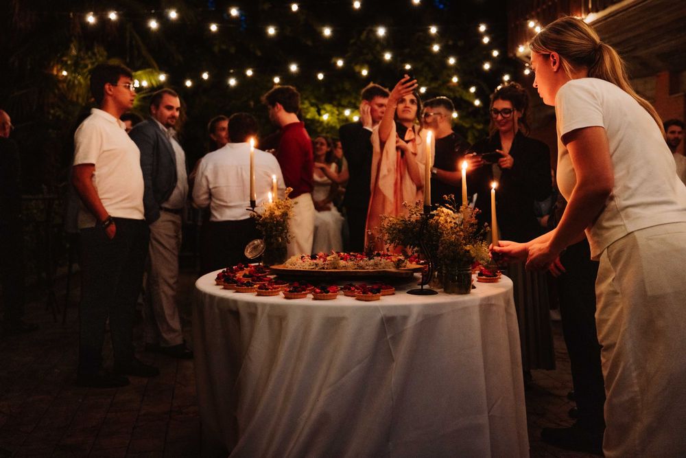 Guests gather around a candlelit table at an outdoor evening celebration with string lights overhead.