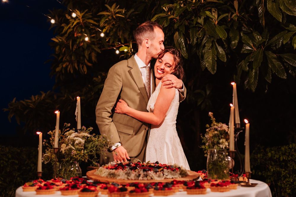 Newlywed couple embracing and kissing beside a floral wedding cake table with candles at an outdoor night reception.