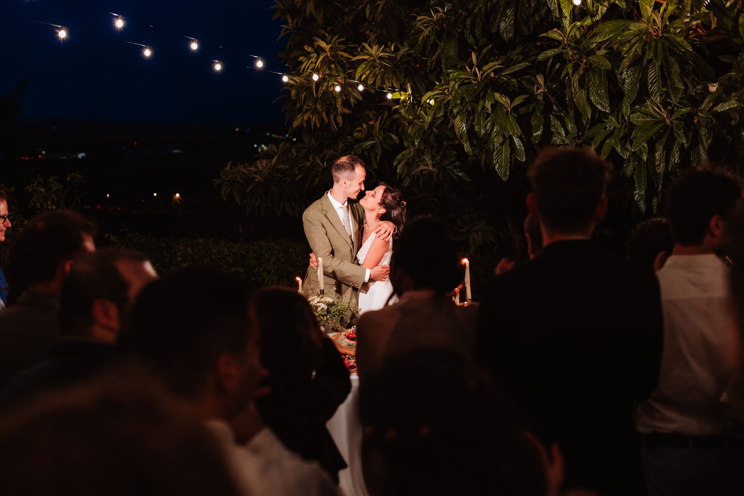 Couple shares romantic first dance under string lights at outdoor evening wedding reception surrounded by guests.
