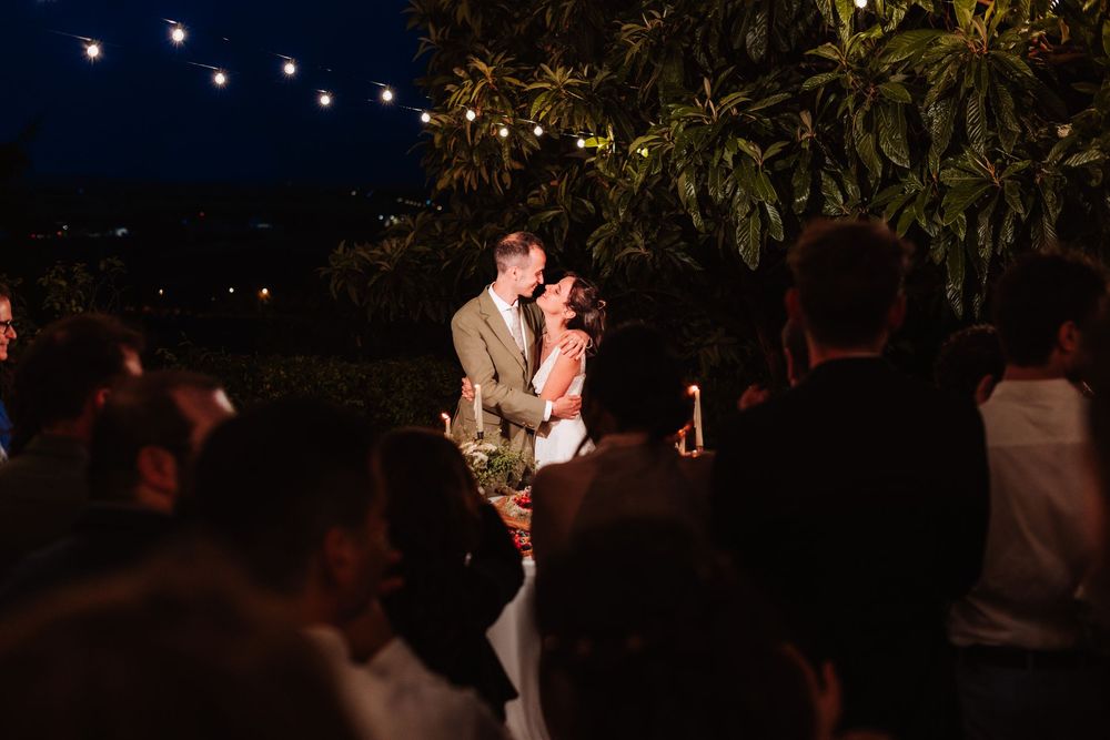 Couple shares romantic first dance under string lights at outdoor evening wedding reception surrounded by guests.