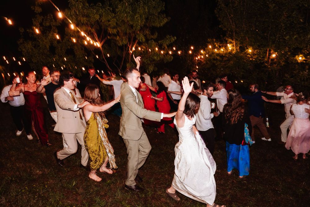 Bride and groom dance with guests at outdoor wedding reception under string lights at night.