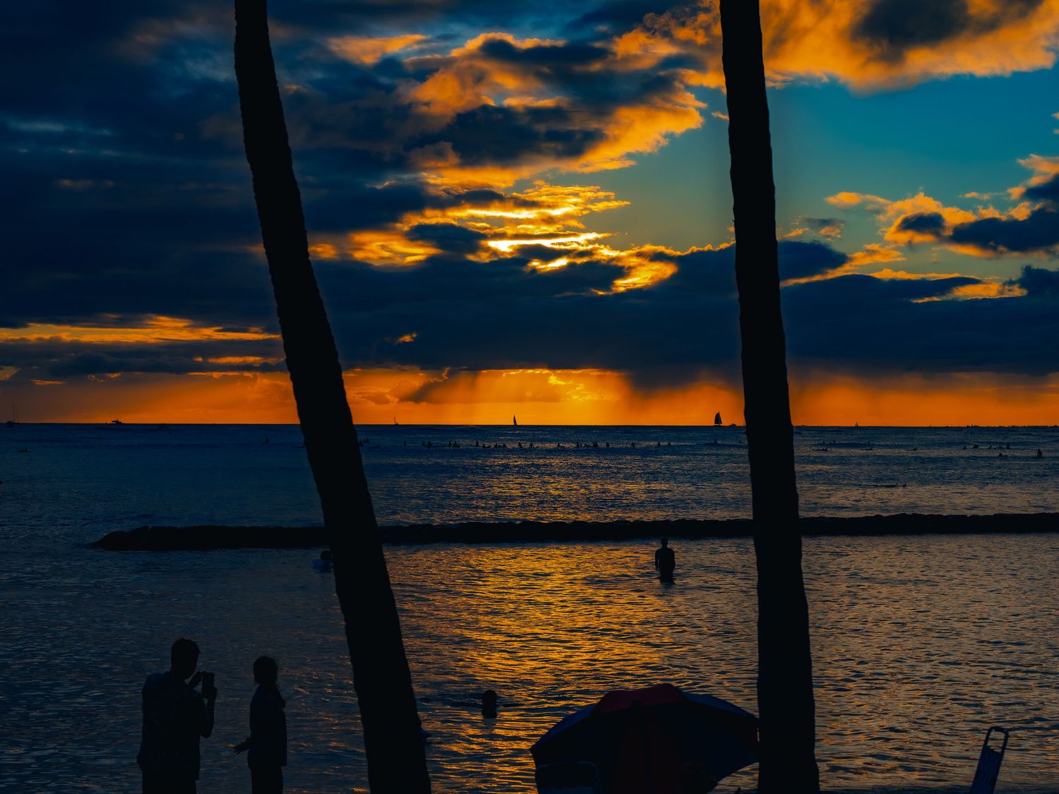 A fiery orange sunset over the ocean with silhouetted palm trunks, sailboats and beachgoers along the shore in Waikiki, Honolulu, Hawaii.