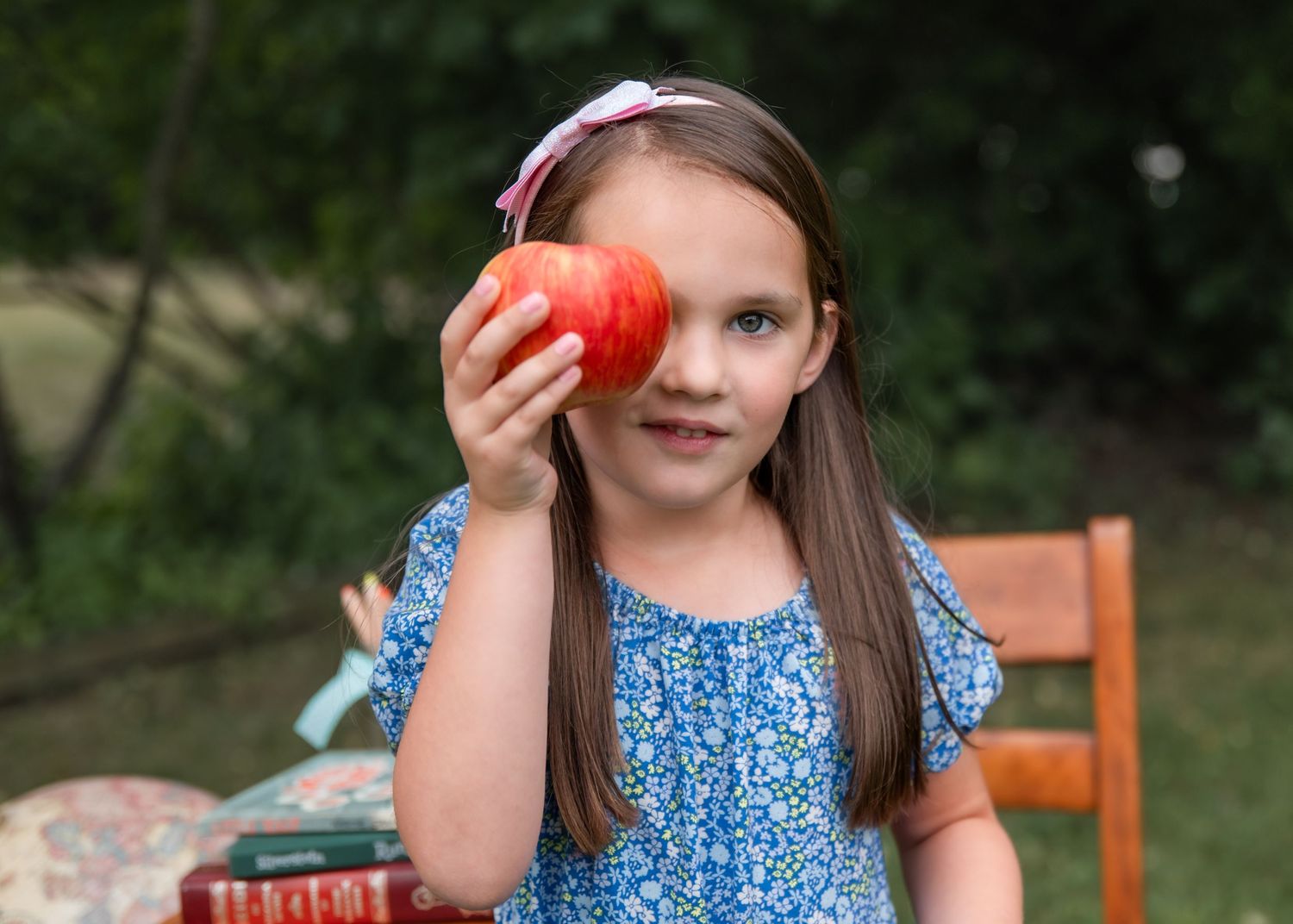 Young girl in blue floral dress holds red apple over one eye, sitting outdoors near books and wooden chair.