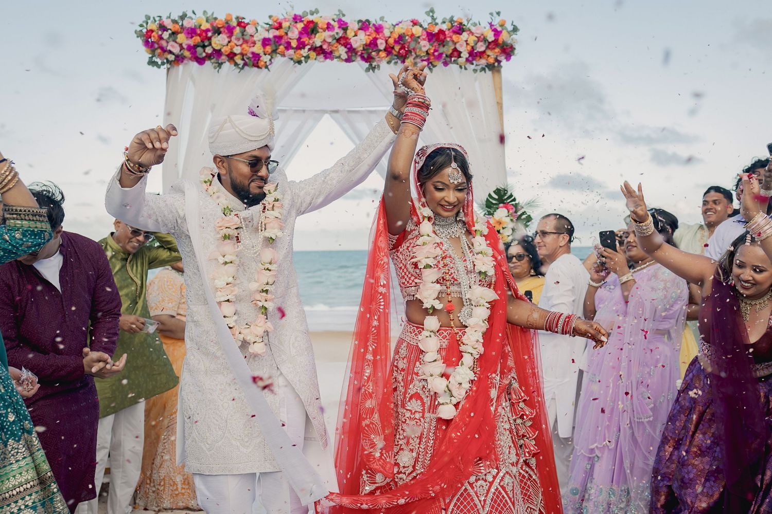 Indian bride in red lehenga and groom in white sherwani celebrate beach wedding ceremony with flower confetti.