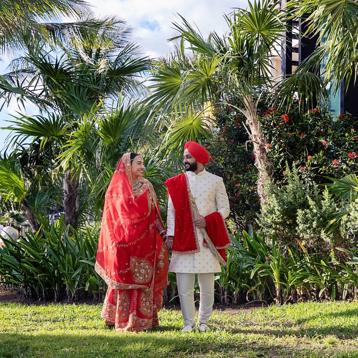Indian bride and groom in traditional red and gold wedding attire standing in a lush tropical garden.