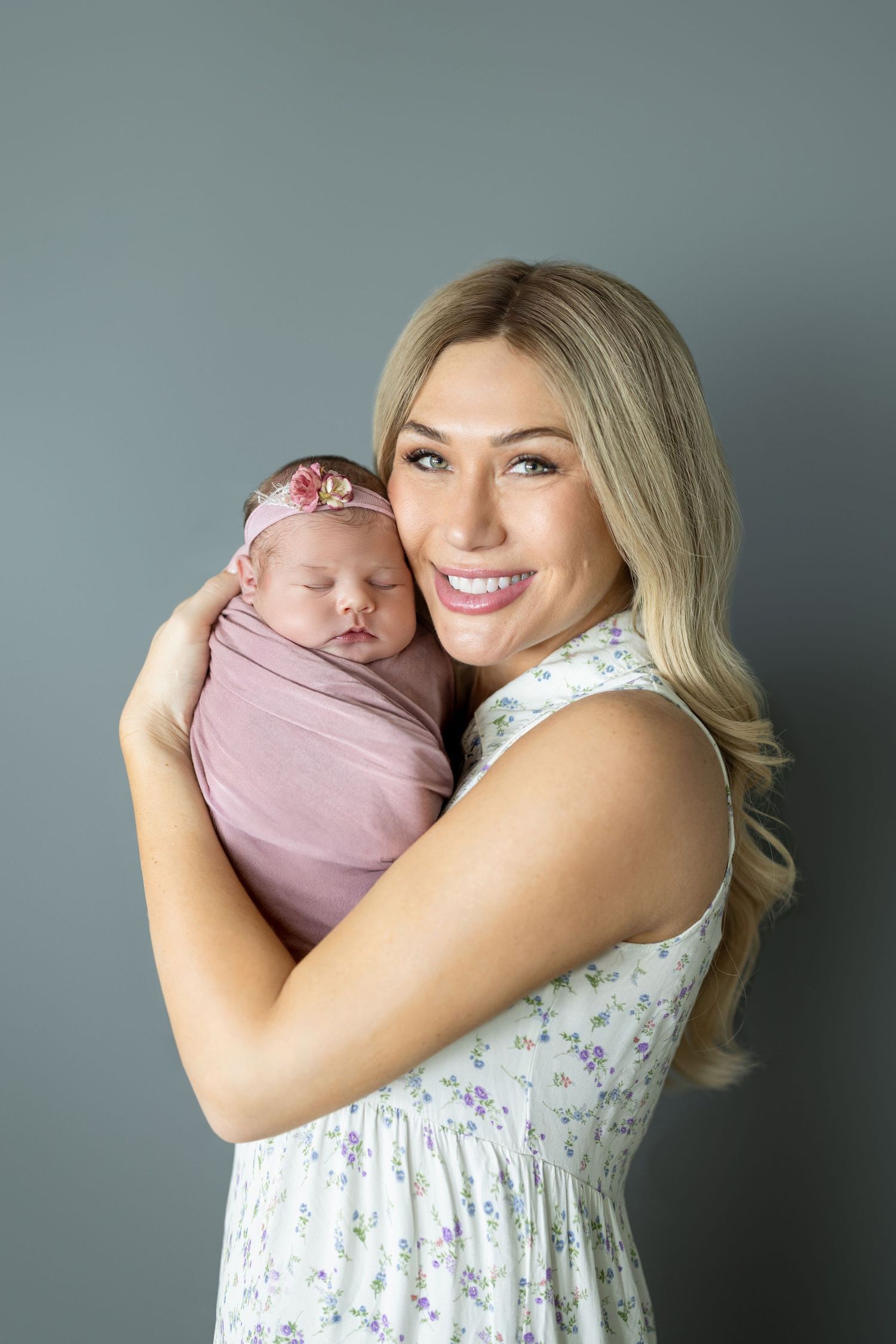 Smiling blonde mother in floral dress holds sleeping newborn baby girl swaddled in pink with floral headband.