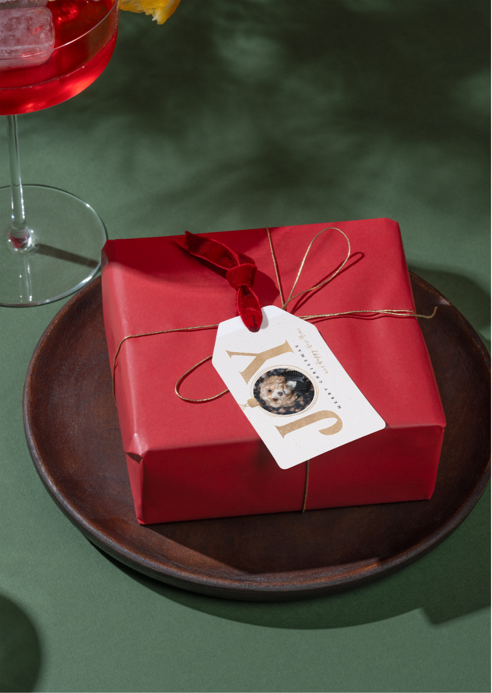 Red gift box with gold twine and tag on dark plate, beside a cocktail glass on green surface.
