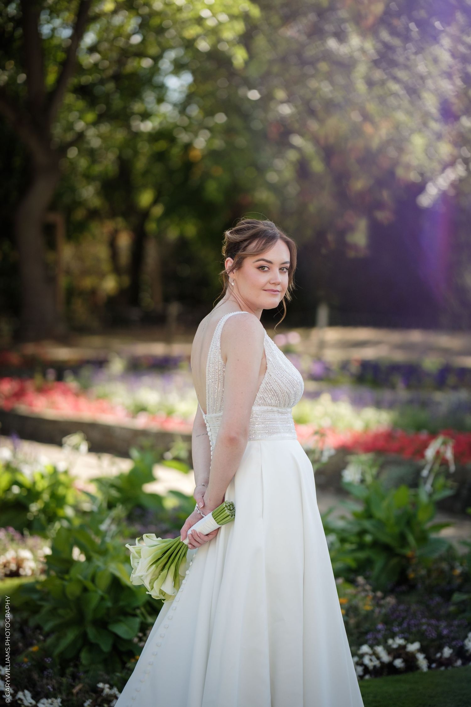 Bride in white gown holding green bouquet poses in sunlit garden with colorful flowers and trees.