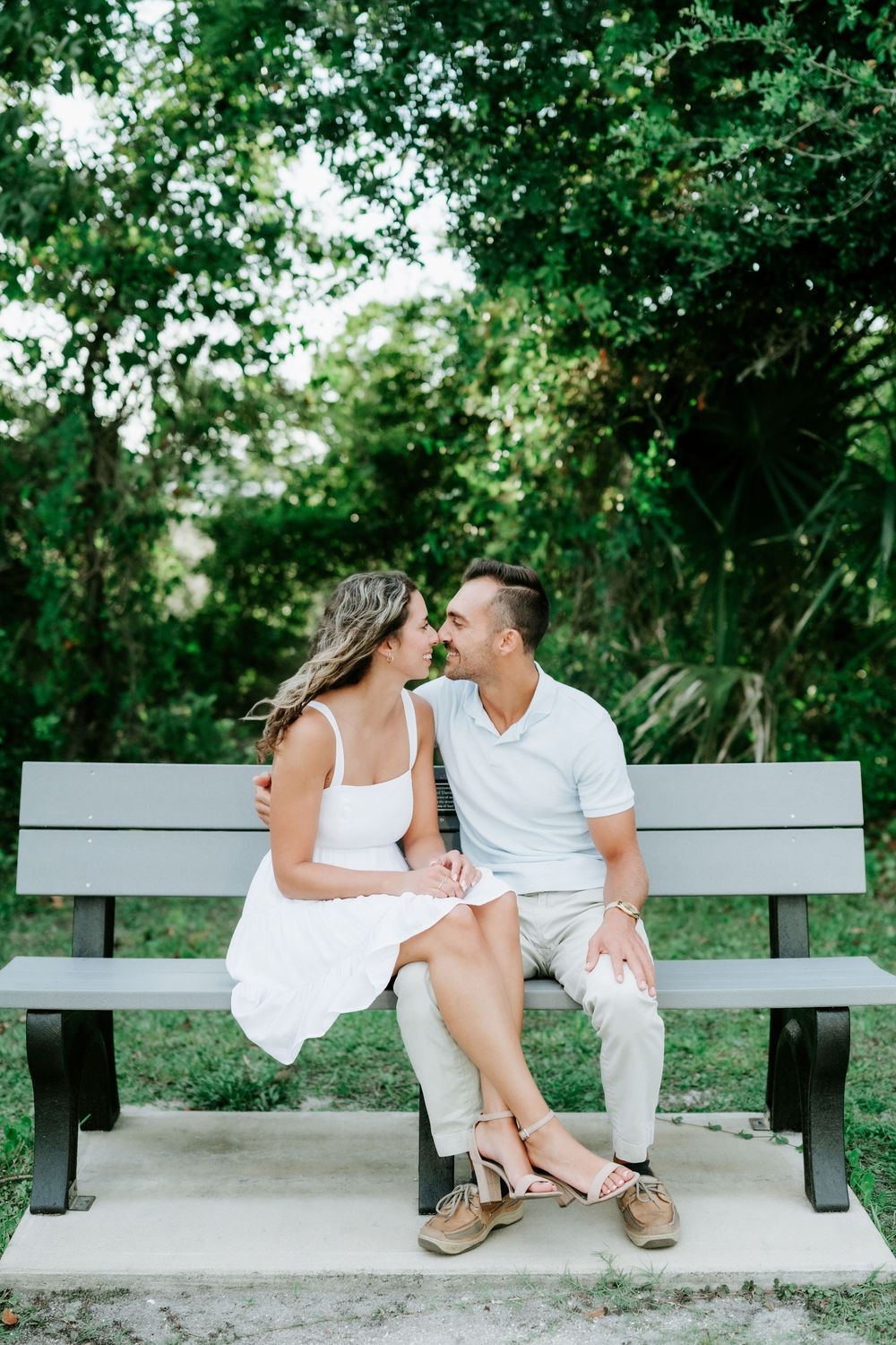 Couple sitting close on a park bench surrounded by lush green trees, wearing white outfits on a sunny day.