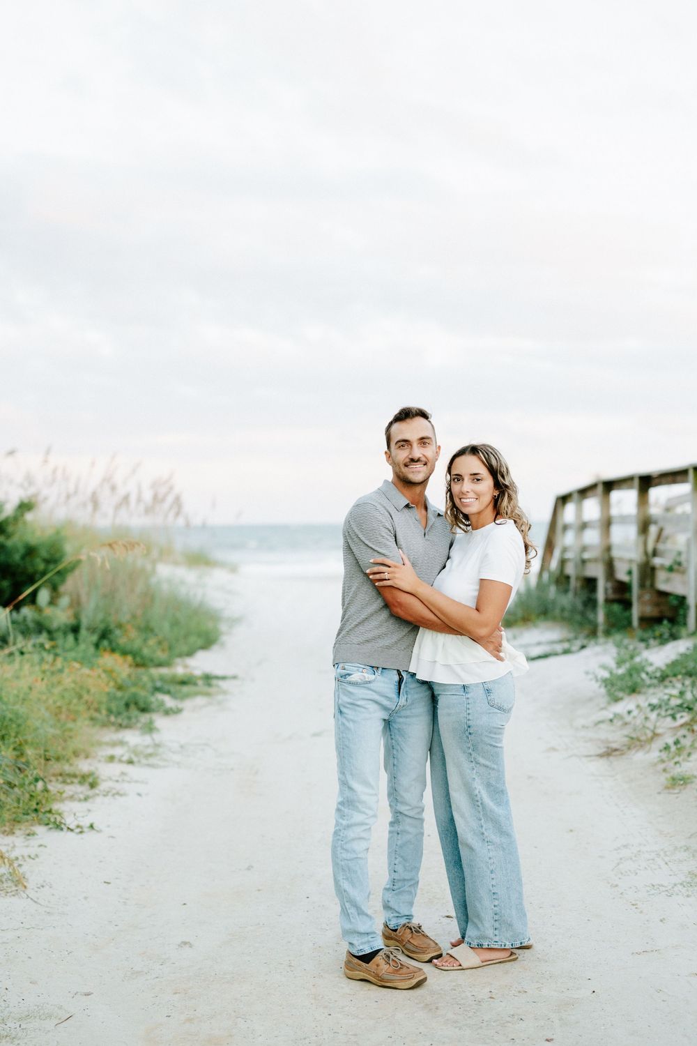 A smiling couple embraces on a sandy beach path, wearing casual jeans and tops, with sea oats and ocean in the background.