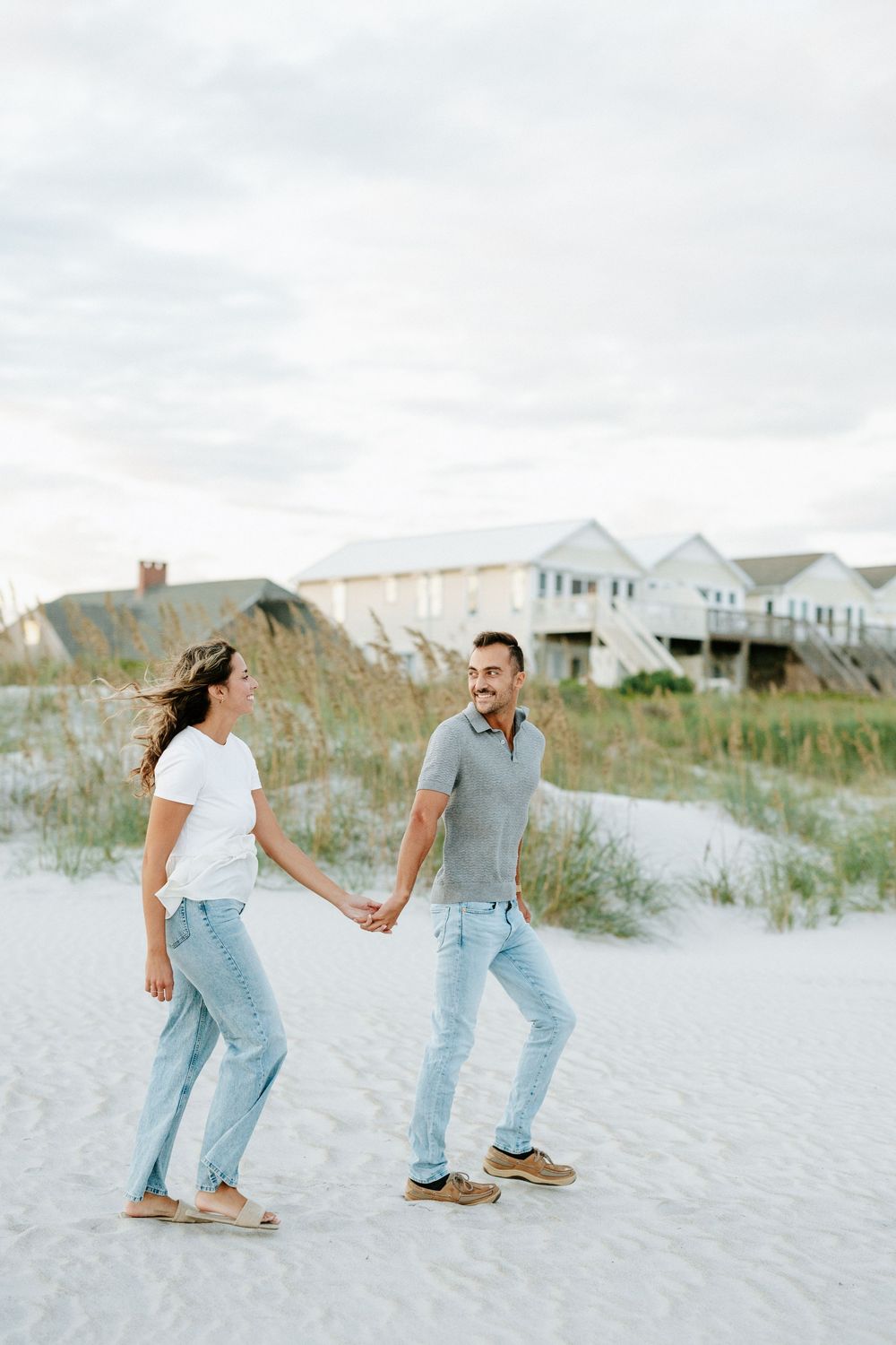 Couple holding hands and smiling on a white sand beach with coastal houses in the background.