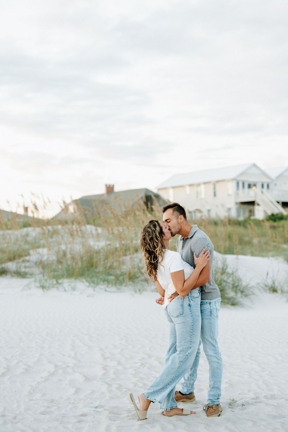 Couple sharing a romantic kiss on a white sand beach with sea oats and coastal homes in the background.