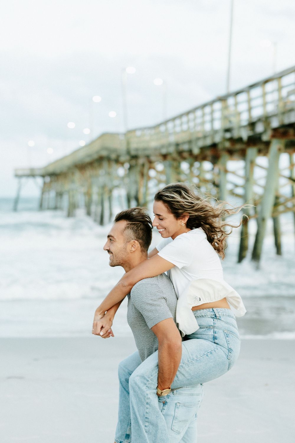 A happy couple enjoys a piggyback ride on the beach near a pier, with the woman smiling and wind blowing her hair.