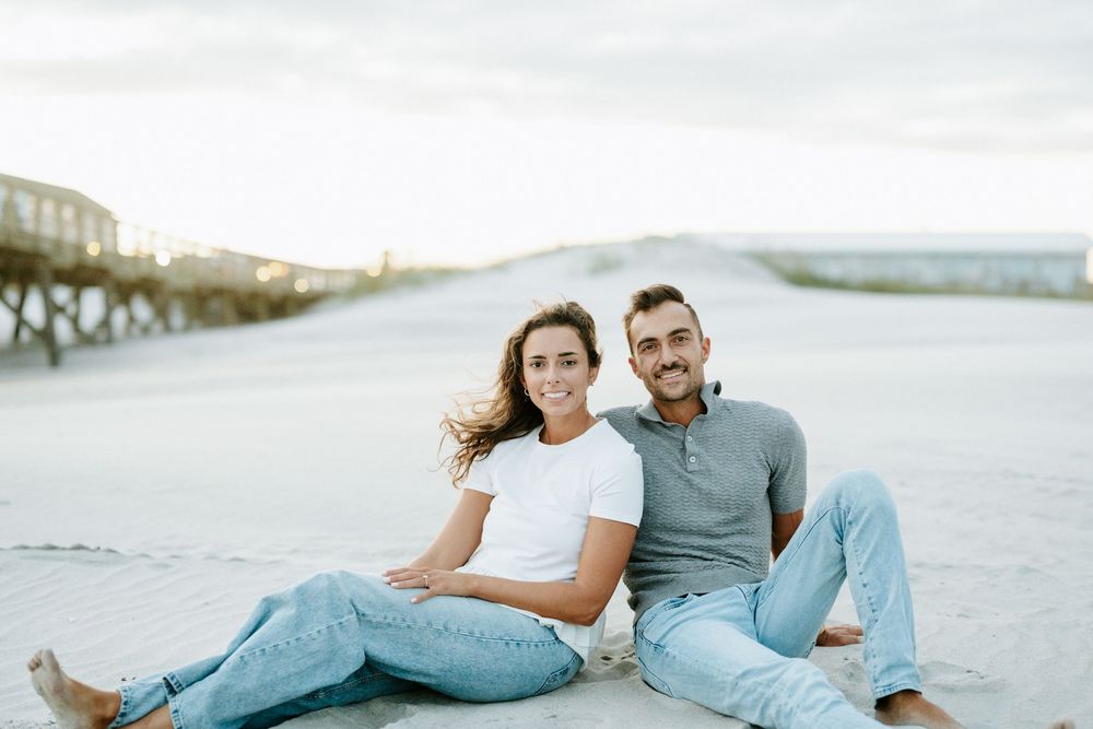 A smiling couple sits together on a sandy beach, wearing casual jeans and light tops, with a pier visible in the background.