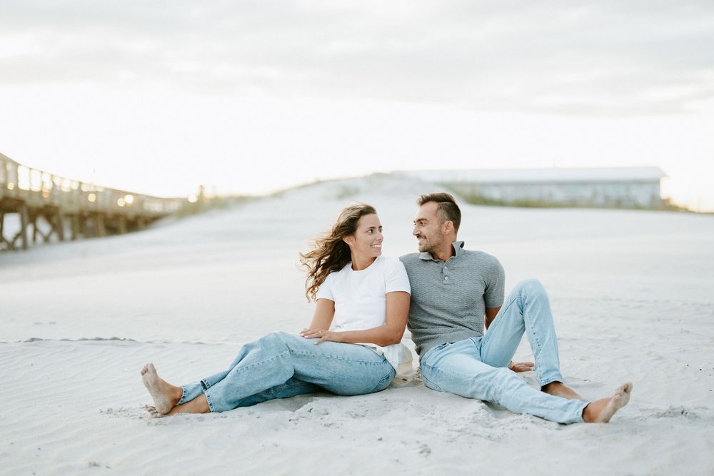 Couple sitting together on white sand beach, smiling at each other, wearing casual jeans and tops with ocean backdrop.