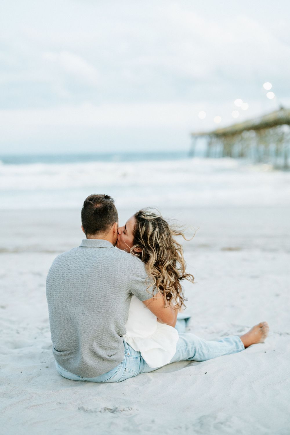 Couple sitting on sandy beach kissing near pier, woman with long wavy hair, both wearing casual jeans and sweaters.