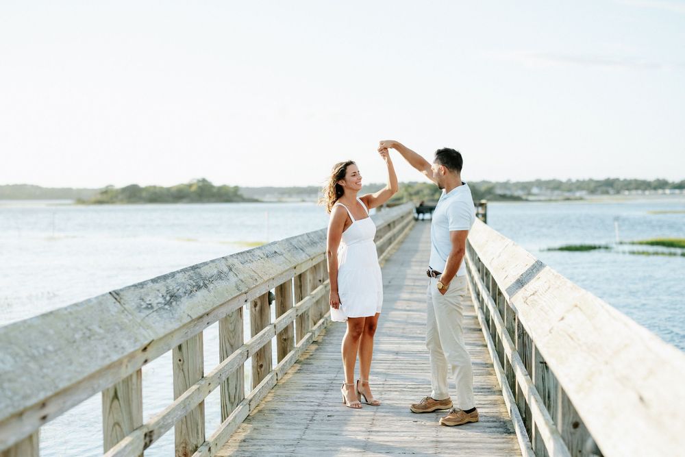 Couple dancing on a wooden pier overlooking calm water, woman in white dress, man in casual attire, sunny day.