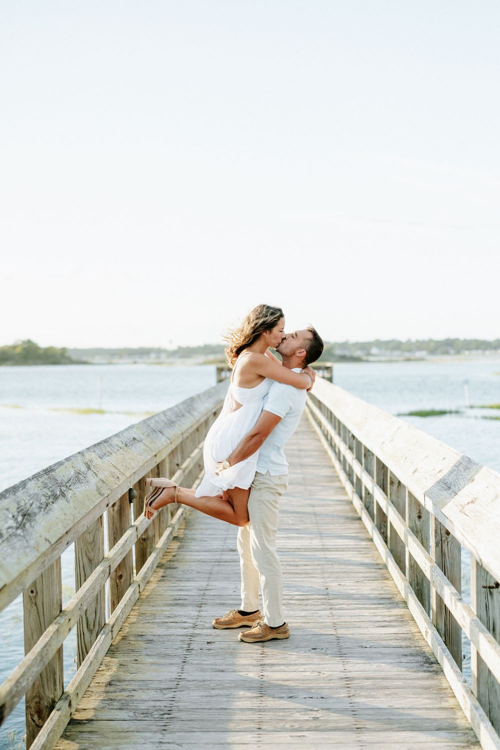 Couple sharing a romantic kiss on a wooden pier over calm water, man lifting woman in white dress.